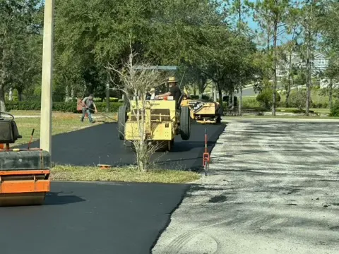 Roller and asphalt paver working together on a wide paving run under trees