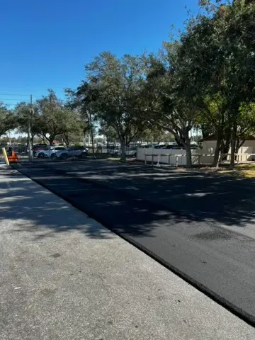 Newly overlaid asphalt roadway running past trees and a fence line
